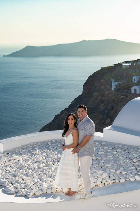 A couple poses happily during a casual photoshoot in Santorini, standing on a rooftop with a breathtaking view of the sea and cliffs in the background.